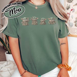 Puede incluir: Una camiseta de manga corta en color verde salvia con un estampado gr&aacute;fico de cuatro peque&ntilde;as criaturas verdes con t&uacute;nicas color canela, dispuestas en una fila horizontal en el pecho. La prenda tiene cuello redondo y un corte holgado.