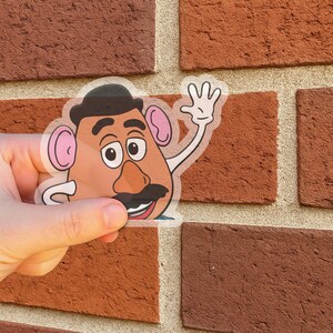 Peut inclure: Un autocollant transparent repr&eacute;sentant un personnage de dessin anim&eacute; avec une t&ecirc;te en forme de pomme de terre, une moustache noire et un chapeau noir. Le personnage fait signe de la main. L'autocollant est tenu devant un mur de briques rouges.