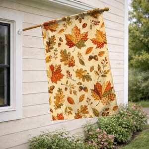 May include: A rectangular garden flag featuring a repeating pattern of autumn leaves, including maple and oak leaves, along with acorns and berries in shades of orange, yellow, brown, and green on a cream-colored background, hanging from a pole.