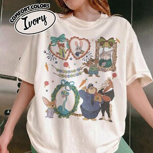 May include: An ivory short-sleeve t-shirt featuring a graphic print of Zootopia characters including Nick, Judy, and others framed in decorative hearts and ovals. The design includes the text Welcome to Zootopia, carrots, and small stars.