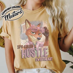 Puede incluir: Una camiseta color amarillo mostaza con un gr&aacute;fico de un zorro de dibujos animados con gafas de sol de persiana rosas y una camisa estampada. El texto dice IT'S CALLED A HUSTLE SWEETHEART. Un logo circular muestra COMFORT COLORS MUSTARD.