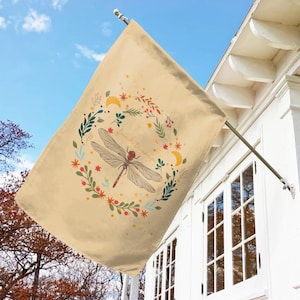 May include: A beige garden flag with a dragonfly and floral design. The dragonfly is red and brown with clear wings. The text "feel the magic" is written in a cursive font.