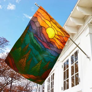May include: A colorful outdoor flag featuring a stained-glass style design of a mountain landscape with a sunset. The flag has a vibrant color palette of orange, yellow, green, and purple. It is mounted on a white house.