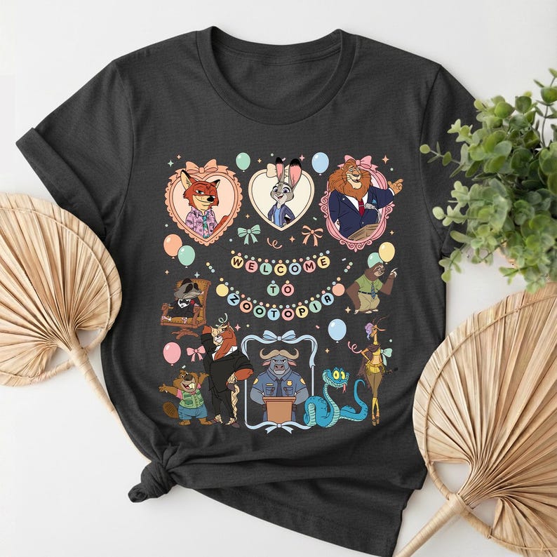 K&ouml;nnte beinhalten: Dunkelgraues T-Shirt mit einem farbenfrohen Aufdruck von Zeichentrickfiguren aus Zootopia. Das Design enth&auml;lt den Text "Welcome to Zootopia" und ist von Herzen, Schleifen und Luftballons umgeben. Ein lustiges, thematisches Design.