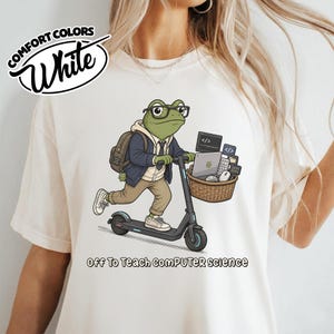 May include: A white t-shirt featuring a graphic of a green frog wearing glasses, a hoodie, and a jacket, riding a black electric scooter. The frog carries a backpack and a basket filled with computer equipment. Text reads: Off To Teach COMPUTER science.