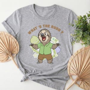 May include: A gray t-shirt featuring a cartoon sloth with the text "WHAT'S THE RUSH?". The sloth is wearing a green shirt and brown shorts, with a surprised expression. The design includes swirls and stars.