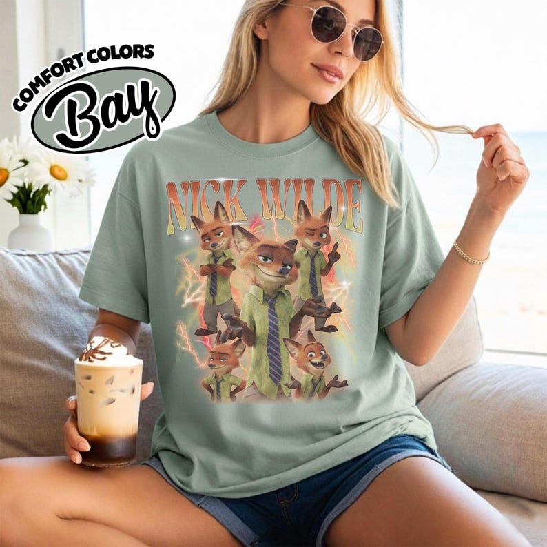May include: A sage green short-sleeve t-shirt featuring a graphic print of the character Nick Wilde in various poses. The text "NICK WILDE" appears in a retro orange font above the character, surrounded by yellow lightning bolt illustrations.