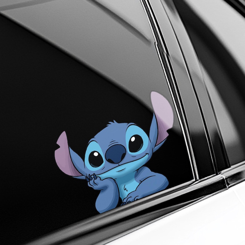 May include: A blue cartoon character sticker, featuring large black eyes and large ears, is affixed to a car window. The character is smiling and resting its chin on its hand. The sticker is a popular character.