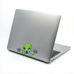 May include: A silver laptop with a colorful sticker of a cartoon character. The sticker features a green hooded figure with a blue face and large eyes. The laptop is open, revealing the keyboard.