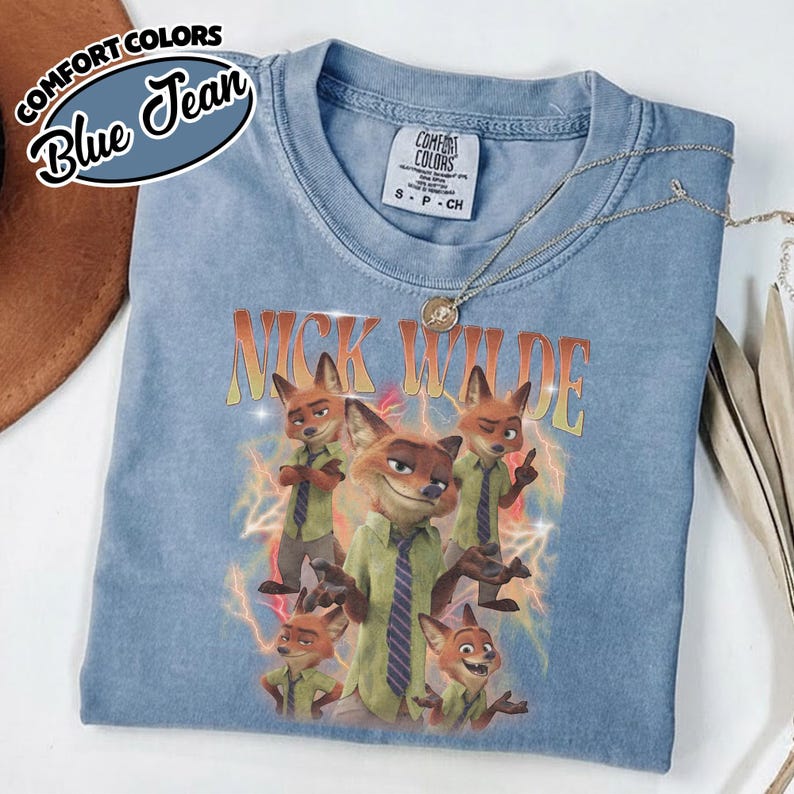 May include: A blue jean colored short-sleeve t-shirt featuring a graphic print of the character Nick Wilde in various poses. The text NICK WILDE appears in a gradient orange font above the character, surrounded by yellow and orange lightning bolts.