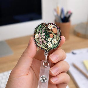 May include: A floral badge reel with a clear plastic strap and a metal clip. The badge reel features a black background with a design of white daisies, purple flowers, and orange accents. The edges have a glittery, iridescent finish.