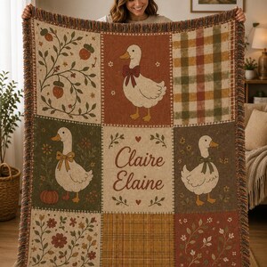 May include: A woven throw blanket featuring a grid of nine panels with illustrations of white ducks wearing bows, acorns, pumpkins, floral patterns, and plaid designs. The center panel displays the text Claire Elaine in brown script.