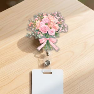 May include: A floral badge reel featuring a bouquet of pink roses, carnations, and other flowers tied with a pink ribbon. The reel has a clear plastic body with a white rectangular card holder.