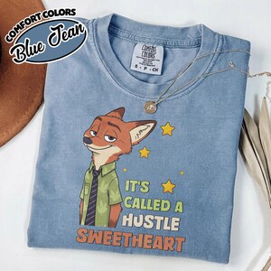 Puede incluir: Una camiseta azul de manga corta con el gr&aacute;fico de un zorro de dibujos animados con camisa verde y corbata. El texto dice IT'S CALLED A HUSTLE SWEETHEART con estrellas amarillas. La camiseta tiene una etiqueta Comfort Colors en talla S.