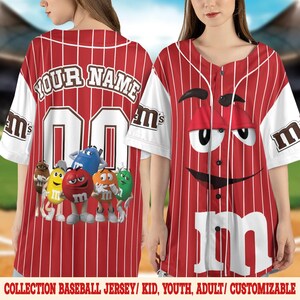 Puede incluir: Camiseta de b&eacute;isbol roja y blanca con rayas verticales. La camiseta tiene el texto "YOUR NAME" sobre el n&uacute;mero "00" en la espalda. La parte delantera muestra un gran dise&ntilde;o de personaje de M&M's. Las mangas son blancas con el logo de M&M's.
