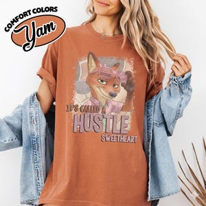 Puede incluir: Una camiseta color &oacute;xido con un gr&aacute;fico de un zorro que lleva gafas de sol de persiana rosas. El texto dice "IT'S CALLED A HUSTLE SWEETHEART". La camiseta tiene cuello redondo y mangas cortas, sobre una camisa vaquera azul claro.