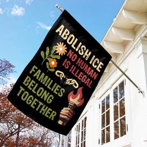 May include: Black flag with the text "ABOLISH ICE, NO HUMAN IS ILLEGAL, FAMILIES BELONG TOGETHER." The flag features a broken chain, flowers, and a torch with a hand holding it. The flag is hanging on a white house.