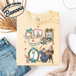 May include: A pale yellow t-shirt featuring a graphic print of Zootopia characters including a fox, rabbit, beaver, snake, and police officer. The design includes heart-shaped frames, a banner reading Welcome to Zootopia, and carrots.