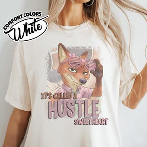 Puede incluir: Una camiseta blanca con el gr&aacute;fico de un zorro que lleva gafas de sol de persiana rosas y una camisa floral. El texto dice IT'S CALLED A HUSTLE SWEETHEART. Un logo en la esquina superior izquierda dice COMFORT COLORS White. Dise&ntilde;o desgastado.