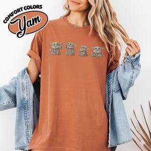 Puede incluir: Una camiseta de manga corta color &oacute;xido con un estampado gr&aacute;fico de cuatro peque&ntilde;as criaturas verdes con t&uacute;nicas beige. El texto COMFORT COLORS Yam aparece impreso en un logotipo ovalado en blanco y negro en la esquina superior izquierda.