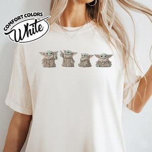 Puede incluir: Una camiseta de color crema con un estampado gr&aacute;fico de cuatro peque&ntilde;as criaturas verdes con orejas y ojos grandes, cada una con una t&uacute;nica de color tostado. El texto COMFORT COLORS White aparece en un logotipo circular negro arriba.