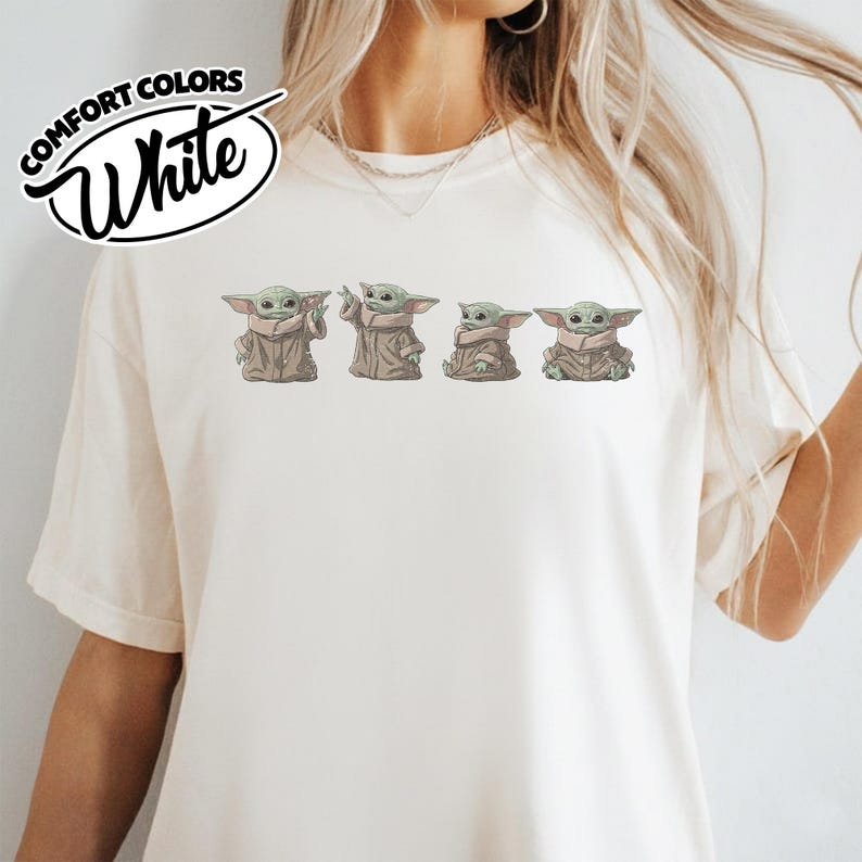 Puede incluir: Una camiseta de color crema con un estampado gr&aacute;fico de cuatro peque&ntilde;as criaturas verdes con orejas y ojos grandes, cada una con una t&uacute;nica de color tostado. El texto COMFORT COLORS White aparece en un logotipo circular negro arriba.