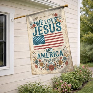 May include: A beige flag with the text "We Love Jesus and America Too" and an American flag design. The flag is decorated with floral accents in red, blue, and yellow. It is hanging on a wooden pole.