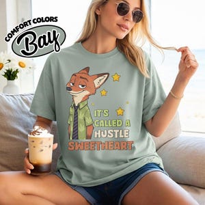 Puede incluir: Una camiseta de manga corta color verde salvia con el gr&aacute;fico de un zorro de dibujos animados con camisa verde y corbata oscura. El texto dice "IT'S CALLED A HUSTLE SWEETHEART" junto a estrellas amarillas. Tiene cuello redondo y corte holgado.