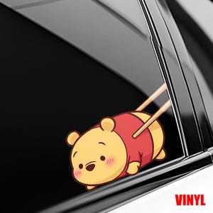 May include: A yellow and red cartoon sticker of Winnie the Pooh being held by chopsticks. The sticker is on a car window. The word "VINYL" is printed in red on the bottom right corner.