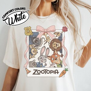 May include: A cream-colored t-shirt featuring a graphic print of Zootopia characters in a polaroid frame. The design includes a pink polka-dot bow, a ZPD badge, a traffic cone, a carrot, and the text Zootopia. The shirt has a crew neckline.