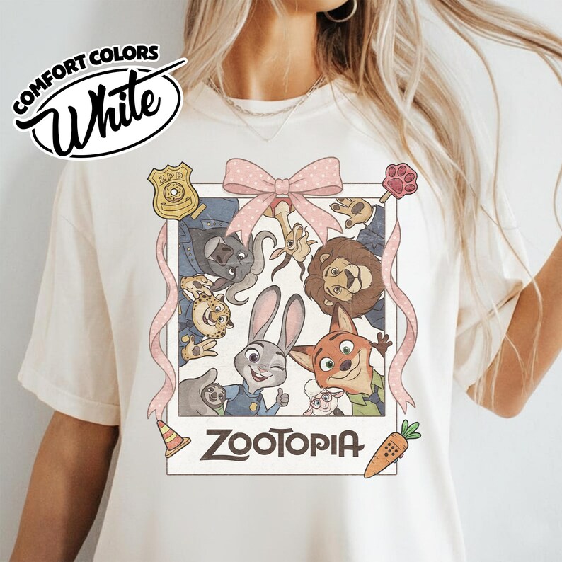 May include: A cream-colored t-shirt featuring a graphic print of Zootopia characters in a polaroid frame. The design includes a pink polka-dot bow, a ZPD badge, a traffic cone, a carrot, and the text Zootopia. The shirt has a crew neckline.