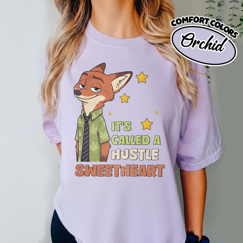 Puede incluir: Una camiseta de manga corta de color morado claro con un gr&aacute;fico de un zorro de dibujos animados con camisa verde y corbata oscura. El texto dice IT'S CALLED A HUSTLE SWEETHEART, junto a peque&ntilde;as ilustraciones de estrellas amarillas. Etiqueta Comfort Colors Orchid.