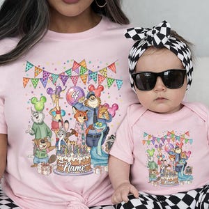 May include: Pink t-shirt and baby bodysuit set featuring a colorful cartoon illustration of animal characters, a birthday cake, and festive bunting. The t-shirt is worn by an adult, and the bodysuit is worn by a baby.