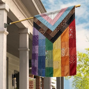 May include: A colorful Pride flag made of patchwork fabric hangs from a wooden pole. The flag features a chevron design at the top with pink, light blue, and brown patches. The lower portion displays a rainbow of colors.