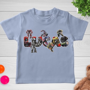 May include: A light blue t-shirt featuring the name "LUCAS" in large, colorful letters. The letters are filled with images of robots. The shirt is a short-sleeved crew neck.