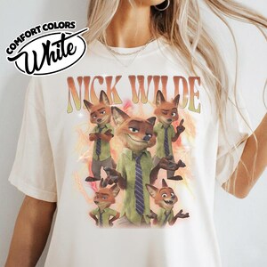 May include: A white t-shirt featuring a graphic print of the character Nick Wilde from Zootopia in various poses. The text "NICK WILDE" appears in a large, stylized orange and yellow font above the character illustrations, set against a background of orange lightning bolts.