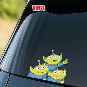 May include: A vinyl decal featuring three green cartoon aliens with three eyes, wearing blue uniforms. The decal has the word "VINYL" in red block letters at the top.