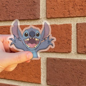 May include: A transparent sticker featuring the character Stitch from the Disney movie Lilo & Stitch. The sticker shows Stitch with a wide open mouth, arms outstretched, and a blue and gray color scheme. The sticker is held in front of a brick wall.