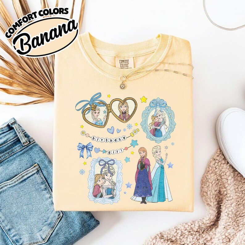 May include: A pale yellow t-shirt featuring a graphic print of two animated characters in heart-shaped lockets, ornate frames, and a banner with the text "birthday girl". The design includes blue bows, stars, and snowflakes, with the characters standing together.