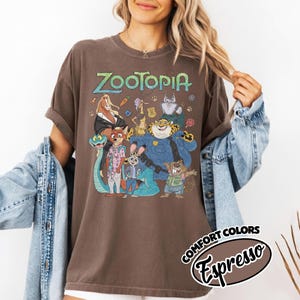 May include: A brown short-sleeve t-shirt featuring a graphic print of Zootopia characters including Nick Wilde, Judy Hopps, and Chief Bogo. The text Zootopia is printed in a light green font above the characters. The shirt is labeled as Comfort Colors Espresso.