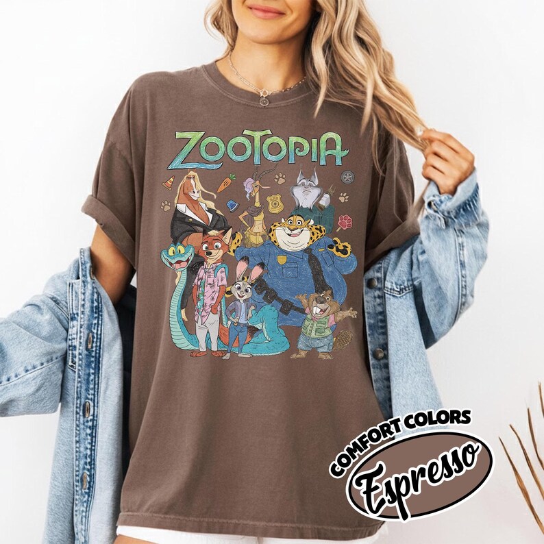 May include: A brown short-sleeve t-shirt featuring a graphic print of Zootopia characters including Nick Wilde, Judy Hopps, and Chief Bogo. The text Zootopia is printed in a light green font above the characters. The shirt is labeled as Comfort Colors Espresso.