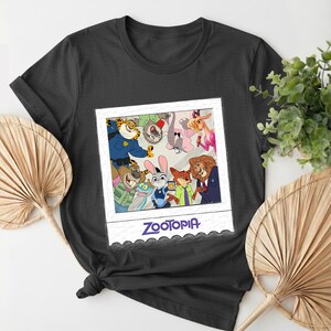 May include: A dark gray t-shirt featuring a Zootopia-themed graphic. The graphic is a polaroid-style image with various cartoon characters from the movie. The word "Zootopia" is printed in blue at the bottom of the image.