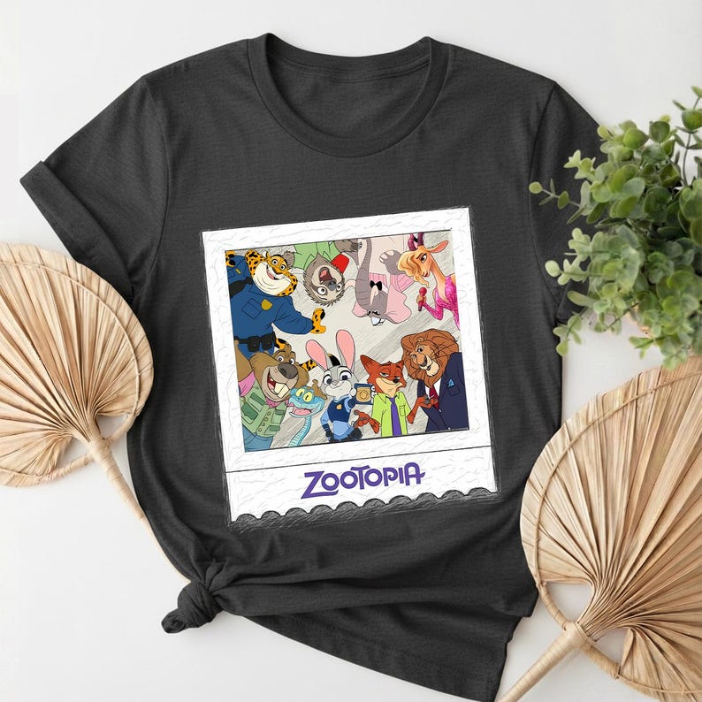 May include: A dark gray t-shirt featuring a Zootopia-themed graphic. The graphic is a polaroid-style image with various cartoon characters from the movie. The word "Zootopia" is printed in blue at the bottom of the image.