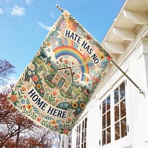 May include: A decorative garden flag with the text "HATE HAS NO HOME HERE" and "HOME HERE". The flag features a colorful illustration of a house, rainbow, flowers, and butterflies. The flag is hanging on a white house.