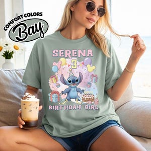 Peut inclure: Un t-shirt vert sauge avec une illustration du personnage Stitch portant un chapeau de f&ecirc;te, entour&eacute; de ballons, de cadeaux et d'un g&acirc;teau d'anniversaire. Le texte indique SERENA 3 BIRTHDAY GIRL. Le t-shirt a un col rond et des manches courtes.