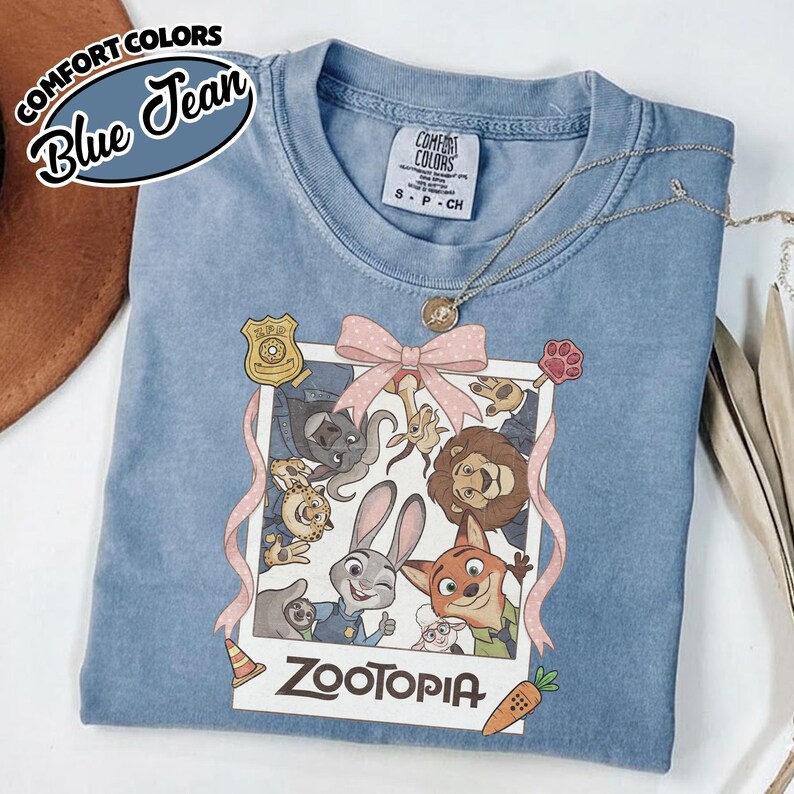 May include: A blue jean colored t-shirt featuring a graphic print of Zootopia characters inside a polaroid frame with a pink bow. The print includes a ZPD badge, a paw print, a traffic cone, and a carrot. The text Zootopia is printed below.