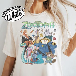 May include: A cream-colored t-shirt featuring a graphic print of Zootopia characters including a fox, rabbit, cheetah, and others. The text "ZOOTOPIA" appears at the top in green, and a "COMFORT COLORS White" logo is visible in the upper left corner.