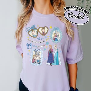 May include: A light purple t-shirt featuring a graphic print of characters from Frozen. The design includes heart-shaped lockets, ornate frames, blue bows, and the text "birthday girl" on a string of beads. The shirt is labeled Comfort Colors Orchid.