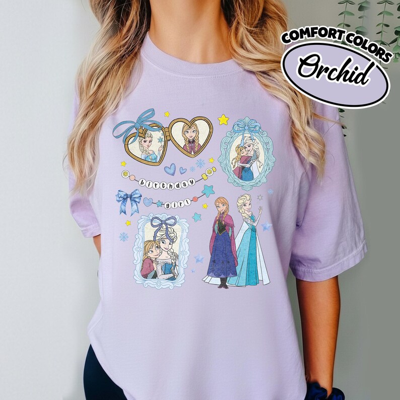May include: A light purple t-shirt featuring a graphic print of characters from Frozen. The design includes heart-shaped lockets, ornate frames, blue bows, and the text "birthday girl" on a string of beads. The shirt is labeled Comfort Colors Orchid.