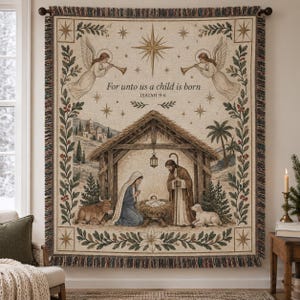 May include: A woven tapestry throw blanket featuring a nativity scene with a manger, figures, and animals. The text reads For unto us a child is born ISAIAH 9:6. The design includes angels, stars, and botanical borders in muted earth tones.